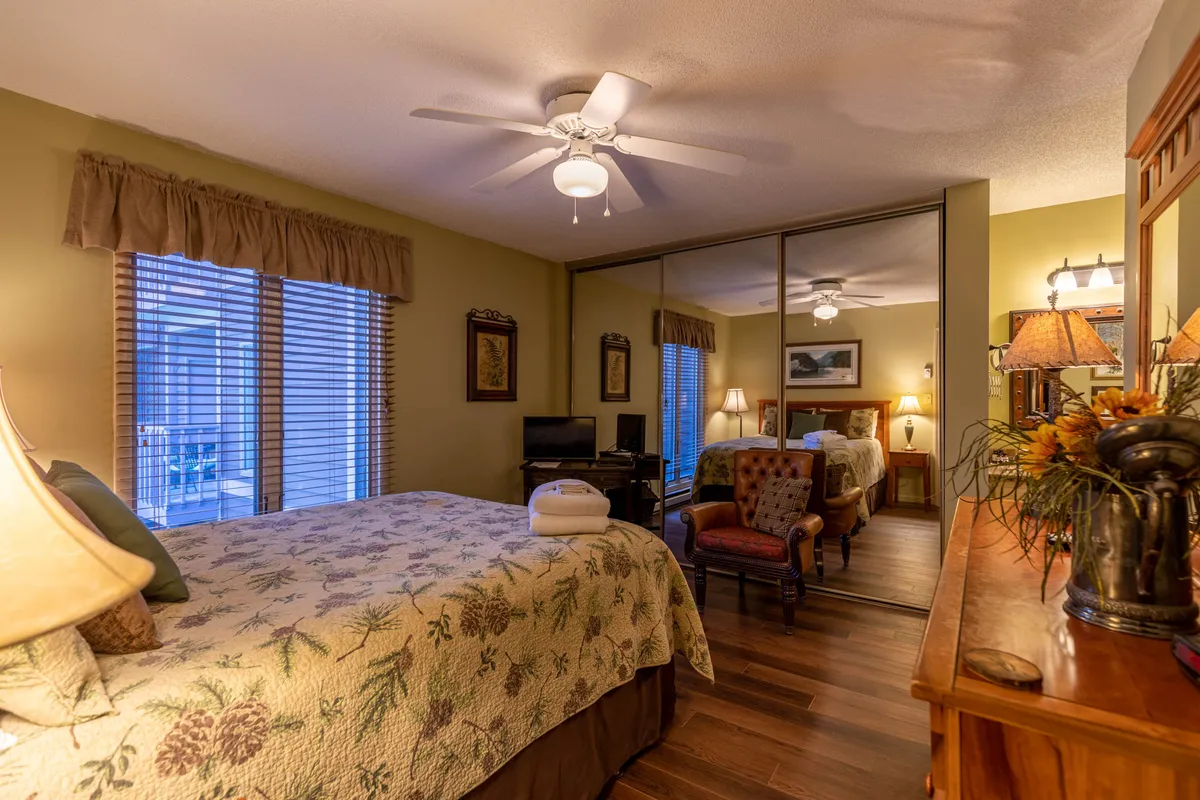 This inviting bedroom features a queen bed adorned with a floral quilt, a cozy armchair set beside decorative lamps, and a mirrored closet that enhances the sense of space. The overall mood is calm and comfortable, perfect for relaxation or enjoying a quiet moment with a book or coffee.