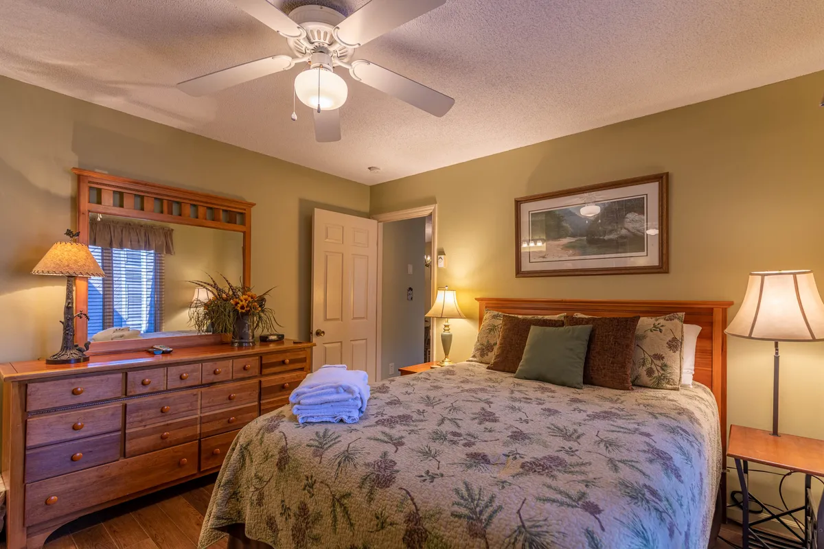 A serene bedroom setting with a wooden queen bed frame, decorative quilt, and matching nightstands topped with lamps. A dresser with a mirror and tasteful artwork create a sense of personalized comfort and style, ideal for restful nights and gentle morning wake-ups.