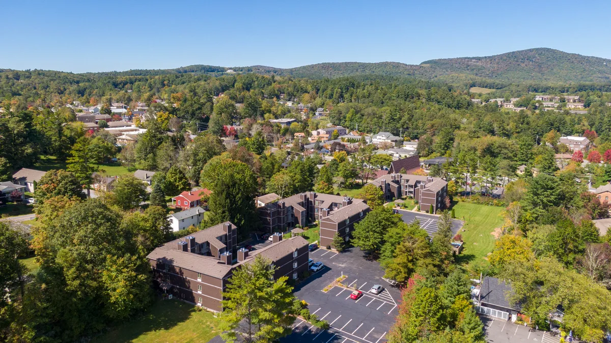 A wide exterior shot featuring residential buildings nestled among greenery with a scenic mountain backdrop. The tranquil setting hints at peaceful evenings and proximity to both natural beauty and town attractions.